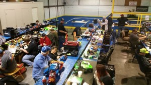 Racers work at pit tables with full electricity and overhead lighting. The onroad track is visible in the back.